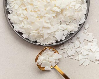 May include: White wax flakes in a gray bowl and a gold spoon. The wax flakes are scattered on a white surface.