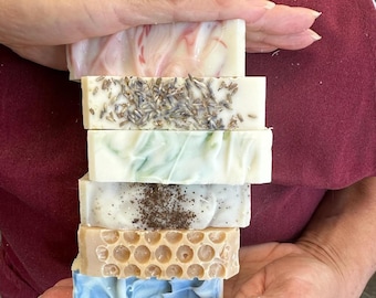 May include: A stack of six handmade soap bars in various colors and textures. The soaps include a marbled pink and white bar, a lavender-topped bar, a green-swirled bar, a speckled bar, a honeycomb-patterned bar, and a blue-swirled bar.