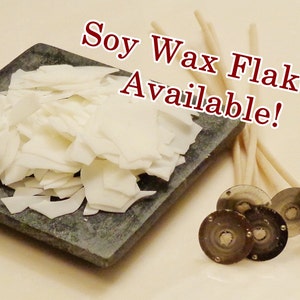 May include: White soy wax flakes on a gray stone surface, next to a bundle of white candle wicks and four silver candle wick stickers. The text "Soy Wax Flakes Available!" is written in red letters on a white background.