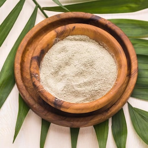 May include: A top-down view of two wooden bowls, one nested inside the other, filled with a light green powder. The bowls are surrounded by green palm leaves. The wood grain is visible on the bowls.