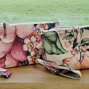 May include: Two floral print cosmetic bags with wrist straps. One bag features large pink and white flowers, while the other has peach and green floral patterns. Both bags have a zipper closure and are made of fabric.