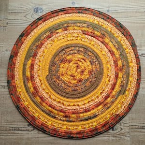 May include: A circular fabric mat constructed from coiled strips of quilted material. The design features concentric rings in warm autumn tones including mustard yellow, burnt orange, deep brown, and olive green with floral patterns.