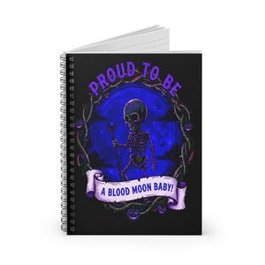 Pode incluir: Caderno espiralado com capa preta com ilustração de esqueleto. O esqueleto, segurando uma bengala, está situado em um fundo de lua azul. O texto "PROUD TO BE" e "A BLOOD MOON BABY!" são exibidos em roxo.