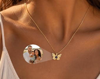Puede incluir: Un collar con colgante de mariposa en tono dorado que presenta una lente de proyección circular central. Las alas de la mariposa están adornadas con pequeñas piedras transparentes. Un inserto circular muestra una foto de un hombre, una mujer y un bebé.