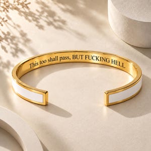 Könnte beinhalten: Ein goldfarbenes Armband in Manschettenform mit einer weißen Emaille-Einlage auf dem äußeren Band. Die Innenseite ist mit dem schwarzen Text graviert: This too shall pass, BUT FUCKING HELL. Das Armband hat ein offenes Design.