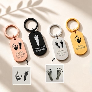 May include: Four metal dog tag keychains in rose gold, black, silver, and gold finishes. Each features engraved handprints or footprints and text including names like Evelyn, Baby Stella, Phoenix, and Saraa, with dates and weights in lbs and oz.