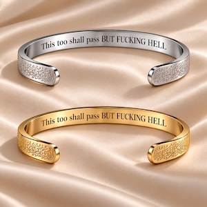 Puede incluir: Dos brazaletes abiertos, uno de tono plateado y otro de tono dorado, cada uno con el texto grabado This too shall pass BUT FUCKING HELL en la superficie interior y patrones decorativos complejos de estilo árabe en los bordes.