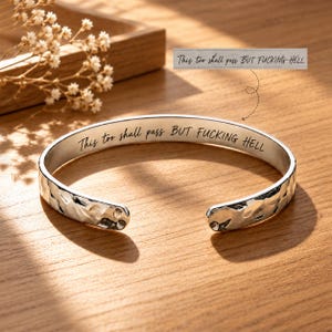 May include: A silver-toned metal cuff bracelet with a hammered, textured exterior. The inner surface is engraved with the black text "This too shall pass BUT FUCKING HELL" in a cursive and sans-serif font combination. The bracelet is open-ended.