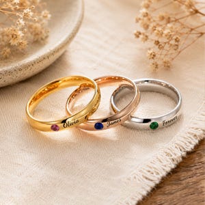 May include: Three metal rings in gold, rose gold, and silver tones. Each ring features a small round gemstone and an engraved name or word. The gold ring has a pink stone and the name Olivia, the rose gold ring has a blue stone and the name James, and the silver ring has a green stone and the word Forever.