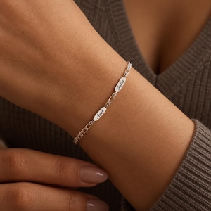 Puede incluir: Una pulsera de cadena en tono plateado que presenta dos pequeñas placas metálicas ovaladas. Cada placa está grabada con un nombre en una fuente cursiva negra, específicamente los nombres Axel y Julia, integrados en los eslabones de la cadena.