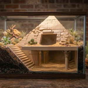 May include: A terrarium with a miniature Egyptian pyramid and desert landscape. The glass enclosure has a dark wood frame and contains sand, plants, and a detailed pyramid structure. The terrarium is lit from above, creating shadows.