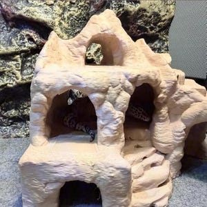 May include: A beige, multi-level reptile hideout featuring a textured rock-like surface, multiple arched openings, and a set of integrated stairs leading to an upper platform. A spotted leopard gecko is partially visible inside the structure.