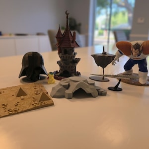 May include: A collection of miniature figurines and models, including a Darth Vader helmet, a castle, a floating island, a character figure, a pyramid model, and a rock formation. The items are arranged on a white surface.