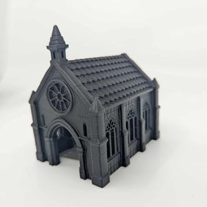 May include: A miniature, dark gray model of a gothic chapel. The chapel features a detailed roof, arched doorway, and a rose window. The structure has multiple arched windows and decorative spires, creating a detailed architectural design.