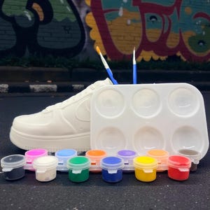 May include: A white sneaker next to a paint palette and small paint pots in various colors. Two paint brushes are in the background. The background features colorful graffiti art.