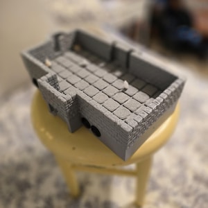 May include: A gray, 3D-printed dungeon terrain piece with a stone brick design. The miniature structure features walls, a floor, and a rectangular shape. The piece is placed on a yellow stool.