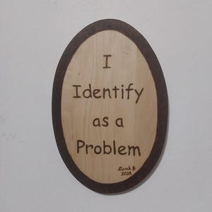Może przedstawiać: Owalna drewniana tablica z ciemnobrązową obwódką. Tablica zawiera tekst "I Identify as a Problem" prostą czcionką. Pod spodem podpis artysty i rok 2025.