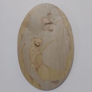 Può includere: Opera d'arte ovale in legno con un design a forma di rana e farfalla. La rana è marrone chiaro con macchie scure e la farfalla è marrone. L'opera è firmata "Linah B. 2024".