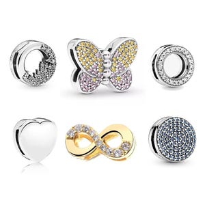 May include: A collection of six metal charms including a butterfly with yellow and pink stones, a smooth silver heart, a gold-tone infinity symbol with clear stones, and three circular charms featuring clear, white, and blue pavé stone settings.