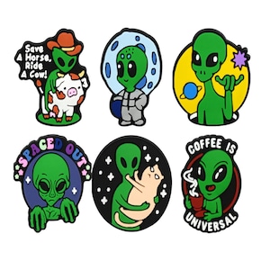 May include: A set of six colourful rubberised pins featuring green alien characters. Designs include an alien with a cow and text "Save A Horse, Ride A Cow!", an astronaut alien, an alien with a planet, "SPACED OUT" text, an alien holding a cat, and an alien with a coffee mug and text "COFFEE IS UNIVERSAL".