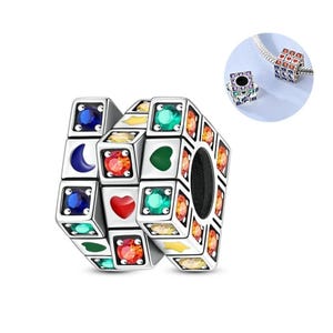 May include: A silver charm with a Rubik's Cube design, featuring colorful gemstones and shapes. The charm includes blue, green, red, and yellow accents, with heart, moon, and square details. A smaller version of the charm is shown in the upper right.