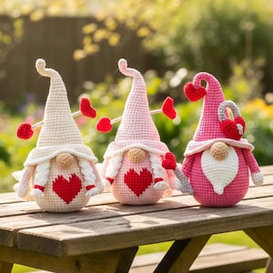 May include: Three crochet gnomes with heart accents sit on a wooden picnic table. The gnomes have white, pink, and red hats, with matching heart-shaped details. Each gnome has a white beard and a red heart on its body. The background is a blurred outdoor scene.