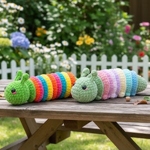 May include: Two crocheted caterpillar toys. One is a vibrant rainbow of colors, the other is pastel. Each has a green head with small antennae and black eyes. They are sitting on a wooden table in a garden setting.