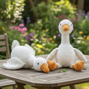 Op de afbeelding: Twee witte gehaakte ganzenknuffels met oranje snavels en poten zitten op een houten tafel. Eén gans staat rechtop, de andere ligt. De achtergrond is een vage tuin met kleurrijke bloemen.