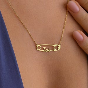 May include: A gold-colored necklace featuring a safety pin design with the word "Lai" in a cursive font. The necklace is worn against a blue background.