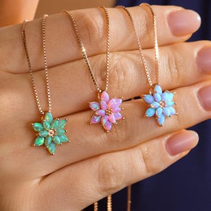 May include: Three gold necklaces with flower pendants in shades of green, pink, and blue. Each pendant features a floral design with multiple petals. The necklaces are held in a hand against a dark blue background.