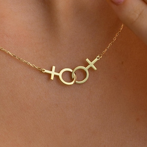 May include: A gold-colored necklace featuring two interlocking Venus symbols. The necklace has a delicate chain and is displayed against a blue background. The Venus symbols are connected by a small ring.