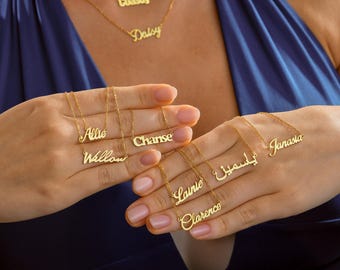 May include: Gold-colored name necklaces with delicate chains. The necklaces spell out names like "Coastly," "Daisy," "Allie," "Willow," "Chanse," "Lainie," "Clarence," and "Janasia." The names are in a cursive font. The necklaces are displayed against a blue background.