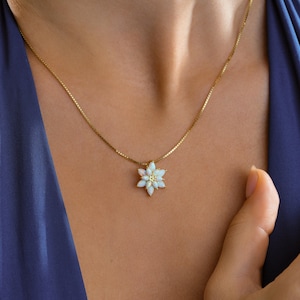 May include: A gold-colored necklace featuring a flower pendant with white petals and a yellow center. The necklace is worn against a blue background, highlighting the jewelry's delicate design. The chain is a fine, square-linked style.