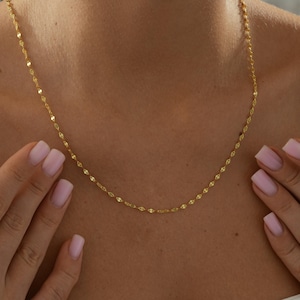 May include: A delicate gold-coloured necklace with a repeating oval link design. The necklace is displayed on a person's chest, with the hands and part of the shoulders visible. The necklace is a simple, elegant piece of jewellery.