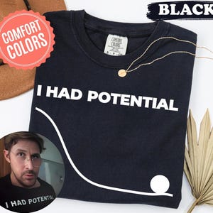 Może przedstawiać: Czarny t-shirt z białym napisem "I HAD POTENTIAL". Widoczny jest również złoty naszyjnik i brązowy kapelusz. Koszulka ma biały wzór graficzny i metkę Comfort Colors.