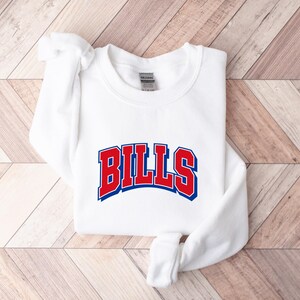 May include: A white long-sleeve crewneck sweatshirt featuring the word BILLS printed in a bold, arched font across the chest. The lettering is red with a thick blue outline and a subtle blue drop shadow, creating a layered, collegiate-style graphic.