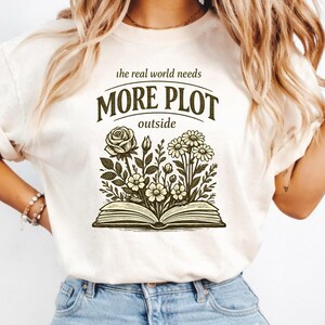 Puede incluir: Una camiseta de manga corta color crema con un gráfico verde oscuro de un libro abierto del que brotan varias flores. El texto sobre la ilustración dice "the real world needs MORE PLOT outside" en una tipografía de color verde oscuro.
