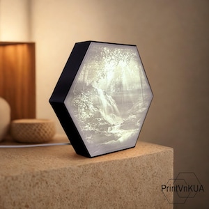 Puede incluir: Lámpara hexagonal negra con una escena de cascada iluminada en su interior. La lámpara está sobre una superficie beige, con un cable blanco en la parte trasera. El diseño de la lámpara es moderno y artístico.