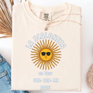 May include: Cream-colored t-shirt featuring the text "LA SCALONETA" in light blue above a yellow sun wearing sunglasses. Below the sun are the words "on Tour" and "USA- CAN- MX 2026" in light blue. The shirt is folded.