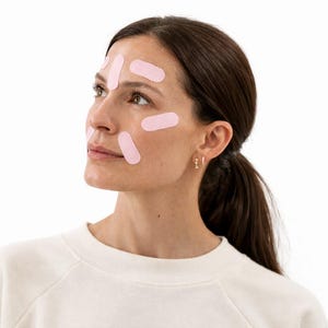 May include: A woman with light pink adhesive strips applied to her face. She is wearing a white long-sleeved shirt and small gold hoop earrings. Her dark hair is pulled back in a low ponytail.