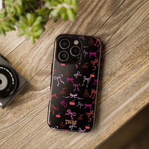 Op de afbeelding: Zwarte telefoonhoes met een patroon van roze en rode kersen en strikken. De hoes heeft een gouden monogram van de initialen "JNM". De telefoonhoes ligt op een houten oppervlak.