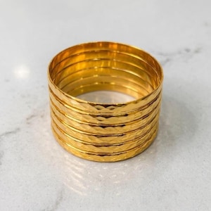 Puede incluir: Un conjunto de siete brazaletes dorados apilados. Cada brazalete presenta una superficie pulida y reflectante con una textura martillada sutil que crea un patrón repetitivo de pequeñas hendiduras poco profundas alrededor de toda la circunferencia.