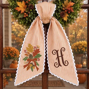 Könnte beinhalten: Ein beigeweiß gestreifter Türhänger aus Stoff mit weißem Wellenrand. Eine Seite zeigt ein gesticktes herbstliches Blumenarrangement mit Blättern, Beeren und Eicheln. Die andere Seite zeigt einen großen, dunkelbraun gestickten Buchstaben H.