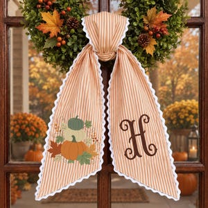 May include: A tan and white striped fabric door hanger with white scalloped trim. One side features an embroidered design of three pumpkins and autumn leaves. The other side displays a large, dark brown embroidered letter H in script font.