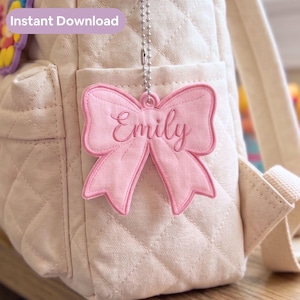 May include: A light pink fabric bow keychain featuring the embroidered name Emily in a cursive script. The bow is attached to a silver-toned metal ball chain and hangs against a quilted cream-colored backpack with an Instant Download label.