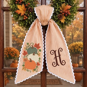 May include: A beige and white striped fabric door hanger featuring a scalloped white trim. One side displays an embroidered design of three pumpkins in green, white, and orange with autumn leaves, while the other side features a large brown letter H.
