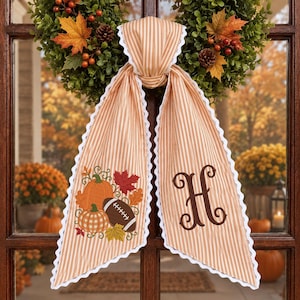 May include: A tan and white striped fabric door hanger with white scalloped trim. One side features an embroidered design of two pumpkins, a football, and autumn leaves. The other side displays a large, dark brown embroidered letter H.