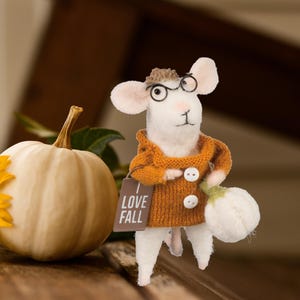 May include: A white mouse figurine wearing a brown sweater, glasses, and a hat, holding a white gourd and a sign that says "LOVE FALL". A white pumpkin is to the left.
