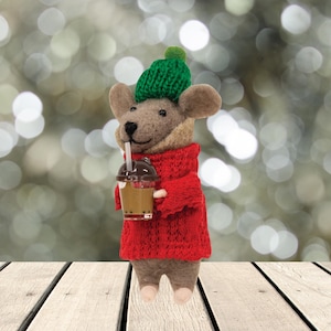 May include: A felt mouse figure wearing a green knitted hat and a red jumper, holding a bubble tea. The mouse has brown fur and is standing on a wooden surface against a blurred background of lights. The figurine is a whimsical holiday decoration.