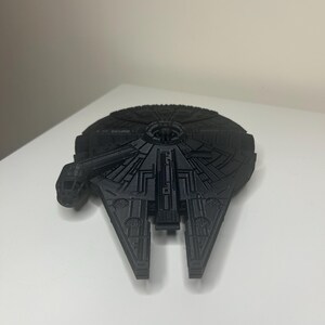 May include: A black model of the Millennium Falcon starship. The model is highly detailed, with visible paneling and design features. The ship's unique shape, including the cockpit and mandibles, is clearly defined. The model is a dark colour.